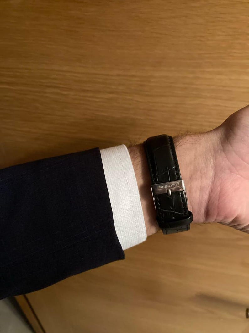 Puede incluir: Un reloj de cuero negro con hebilla plateada abrochado en una mu&ntilde;eca. El reloj se combina con una chaqueta de traje azul marino y un pu&ntilde;o de camisa blanca. La correa del reloj tiene un patr&oacute;n texturizado.