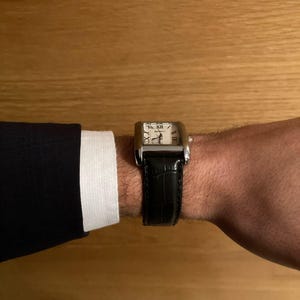 Puede incluir: Un reloj de pulsera rectangular con caja plateada y correa de cuero negro. La esfera del reloj es blanca con n&uacute;meros romanos negros. El reloj est&aacute; en una mu&ntilde;eca, con una chaqueta oscura y un pu&ntilde;o de camisa blanco visibles.