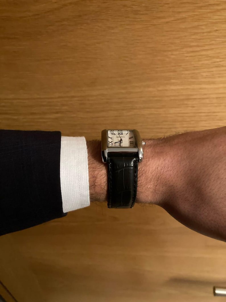 Puede incluir: Un reloj de pulsera rectangular con caja plateada y correa de cuero negro. La esfera del reloj es blanca con n&uacute;meros romanos negros. El reloj est&aacute; en una mu&ntilde;eca, con una chaqueta oscura y un pu&ntilde;o de camisa blanco visibles.