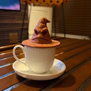 May include: A white teacup and saucer set with a brown Sorting Hat lid. The lid is shaped like the Sorting Hat from the Harry Potter series. The cup and saucer are on a wooden table.
