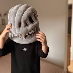 Puede incluir: Una máscara de calavera de felpa gris con dientes grandes y cuencas de los ojos es sostenida por una persona. La persona lleva una camisa negra de manga larga con la palabra "LUC" y un logotipo de calabaza en el pecho.