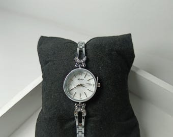 May include: A silver-toned wristwatch with a white face and a delicate bracelet band. The watch features a round face with silver hands and markers. The band has decorative elements with small, clear stones. The watch is displayed on a dark gray pillow.
