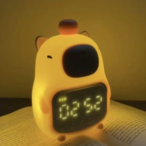 Puede incluir: Un despertador amarillo con forma de oso con una pantalla digital que muestra la hora. El reloj tiene una pantalla negra y está iluminado. Se coloca sobre un libro abierto con texto visible.
