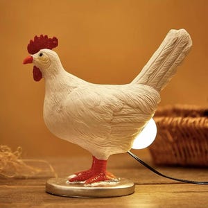May include: A white rooster-shaped lamp with a red comb and feet. The lamp has a round base and a light bulb that shines from the body. The lamp is on a wooden surface.