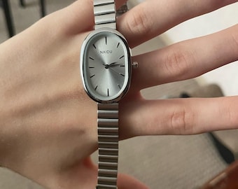 May include: A silver wristwatch with an oval face and a matching metal band. The watch face is silver with black hands and the brand name "NAIDU" printed at the top. The watch is shown against a neutral background.