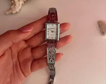May include: A silver-toned quartz watch with a rectangular face and a decorative band. The watch face displays the numbers 1 through 12. The band features an ornate design. The watch is held by a person.