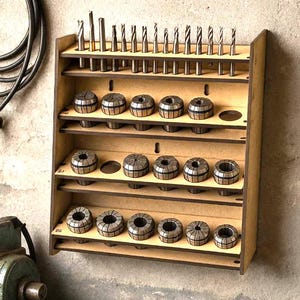 Puede incluir: Un organizador de herramientas de madera con múltiples estantes, que contiene varias brocas y pinzas. Las pinzas están dispuestas en filas y las brocas se almacenan verticalmente. El organizador está montado en una pared texturizada, con un torno visible en primer plano.