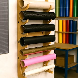 Puede incluir: Un estante de almacenamiento de madera montado en la pared que contiene varios rollos de papel artesanal en varios colores, incluyendo blanco, negro, rosa y dorado. El estante está montado en una pared blanca, con una estera de corte y herramientas a la vista.