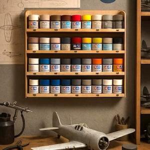 May include: A wooden shelf displays rows of model paint jars in various colors. A gray model airplane sits on a wooden surface with tools and paintbrushes. A drawing of an airplane and a US Air Force emblem are on the wall.