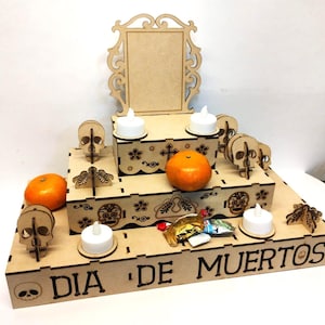 May include: A wooden Dia de Muertos altar with three tiers, adorned with laser-cut skulls, decorative cutouts, and the text "DIA DE MUERTOS". It features two oranges, battery-operated candles, and a decorative frame.