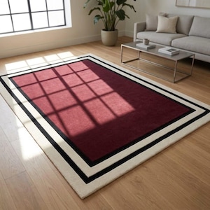May include: A rectangular area rug featuring a solid deep red center panel, framed by a thick black border and an outer cream-colored border. The rug lies flat on a light-toned wooden floor, displaying a smooth, uniform texture across its surface.