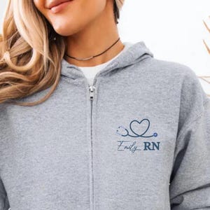 May include: A gray zip-up hoodie with a hood and front pockets. The left chest features a blue stethoscope design with the name "Emily RN" in a script font. The hoodie is made of a soft material.