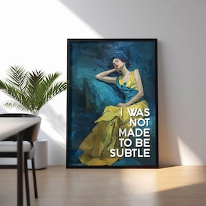 May include: Framed art print depicting a woman in a yellow and blue dress, reclining on a blue surface. The artwork has a painterly style with the text "I WAS NOT MADE TO BE SUBTLE" in white.