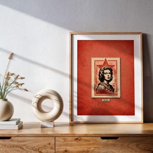 Op de afbeelding: Ingelijste kunstdruk met een vintage postzegelontwerp. De postzegel, tegen een rode achtergrond, toont een vrouwenportret met een ster. Het kunstwerk staat in een houten lijst, tentoongesteld op een houten kast.