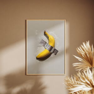 May include: A framed art print featuring a ripe yellow banana duct-taped to a light gray background. The artwork is set against a neutral-toned wall, with a hint of dried floral accents.