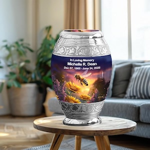 May include: A silver and blue memorial urn with floral and bee imagery. The urn has the text "In Loving Memory Michelle R. Dean Dec 07, 1965 - June 24, 2025". The urn is on a wooden table.