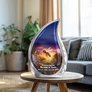 May include: A silver-colored teardrop-shaped urn with a detailed floral design. The urn features a vibrant image of a bee hovering over a sunflower, set against a sunset backdrop. Text reads "In Loving Memory Michelle R. Dean Dec 07, 1965 - June 24, 2025."