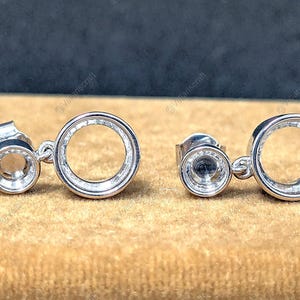 May include: A pair of silver-colored stud earrings. Each earring features a small, round stud connected to a larger, open circle. The earrings are displayed on a tan surface with a dark blue background.