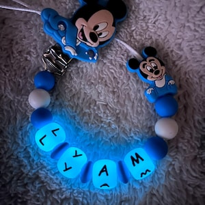 Op de afbeelding: Een blauw-witte fopspeenclip voor baby's met een Mickey Mouse-ontwerp. De clip heeft een reeks blauwe en witte kralen, met oplichtende kralen met letters die een naam spellen. Een Mickey Mouse-figuur is bevestigd.