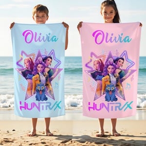 May include: Two beach towels, one light blue and one pink, each featuring a graphic of a star with three female characters, a blue cat, and the name "Olivia" and the word "HUNTRAX". The towels are held by children on a sandy beach.