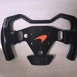 May include: A black steering wheel featuring an orange logo. The wheel has a unique design with curved grips and a central panel. The logo is a stylized orange arrow. The wheel appears to be made of a glossy material.