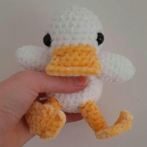 Può includere: Un giocattolo a forma di anatra all'uncinetto fatto a mano. L'anatra ha un corpo, ali e testa bianchi, con becco e zampe gialle. Gli occhi sono neri. Il giocattolo è morbido e soffice ed è tenuto da una persona.