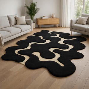 May include: A large, abstract area rug with a black, blob-like design against a cream-colored background. The rug is placed on a hardwood floor in a living room setting, with neutral-toned furniture visible in the background.