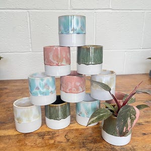 May include: A pyramid stack of ten cylindrical ceramic planters, each featuring a white base and a glazed upper section in shades of blue, pink, green, and cream. One planter contains a small plant with variegated pink and green leaves.
