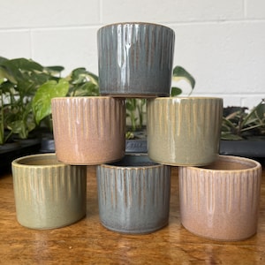 May include: Six small ceramic pots in a stack, in shades of dusty rose, sage green, and muted blue-grey. Each pot features vertical ribbed detailing and a glossy finish, ideal for small plants or succulents.