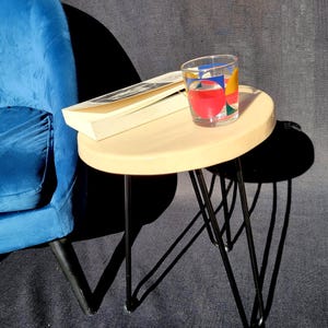 Peut inclure: Petite table d'appoint ronde avec un plateau en bois clair et des pieds en épingle noirs. Un verre avec un motif géométrique coloré et un livre reposent sur la table. Un fauteuil bleu est en arrière-plan.