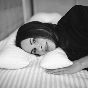 May include: Black and white image of a person lying on a bed with a striped comforter and pillows. The person is wearing a black top and is looking at the camera. The bed has a padded headboard.