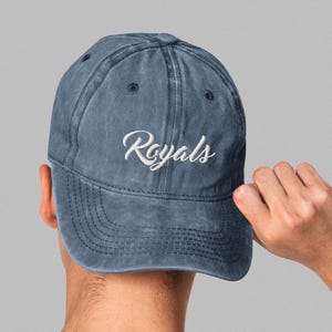May include: A denim blue baseball cap with the word "Royals" embroidered in white script. The cap has a curved brim and a distressed, vintage look. The cap is being held by a person's hand.