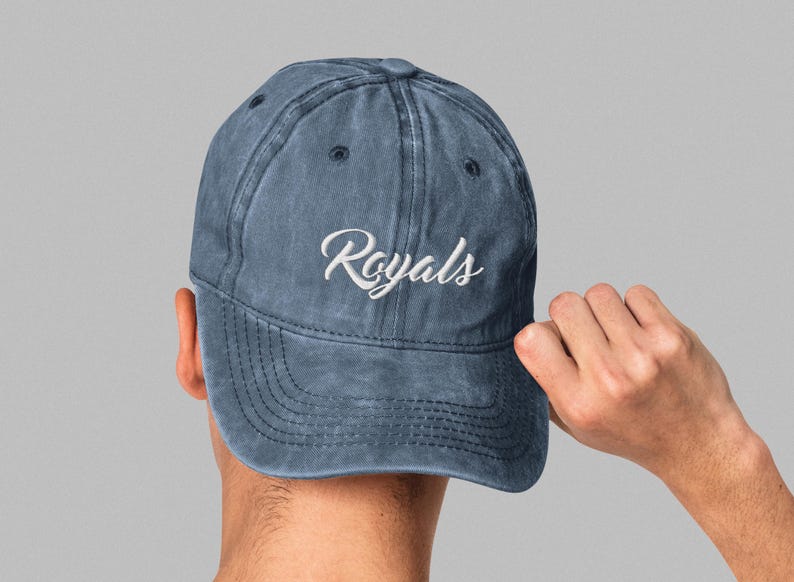 May include: A denim blue baseball cap with the word "Royals" embroidered in white script. The cap has a curved brim and a distressed, vintage look. The cap is being held by a person's hand.