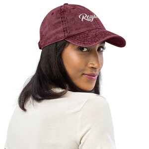 May include: A maroon baseball cap with the word "Royal" embroidered in white script on the front. The cap has a curved brim and an adjustable strap at the back. The cap is made of a soft, worn material.