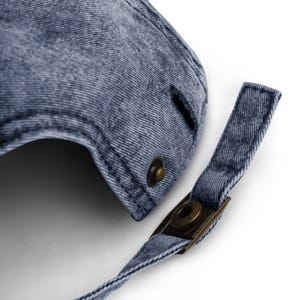May include: Close-up of a denim baseball cap in a faded blue wash. The cap features a curved brim and an adjustable strap with a metal clasp. The metal hardware has a bronze finish. The fabric has a textured appearance.