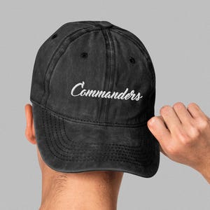 Op de afbeelding: Een zwarte, verweerde baseballcap met het woord "Commanders" in witte letters geborduurd. De pet wordt door een persoon gedragen, met de achterkant van het hoofd en een hand zichtbaar.