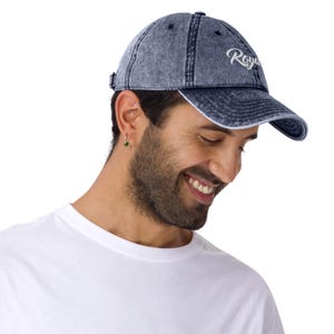 May include: A denim blue baseball cap with the word "Royal" embroidered in white script. The cap has a curved brim and an adjustable strap at the back. The cap is worn by a person wearing a white t-shirt.
