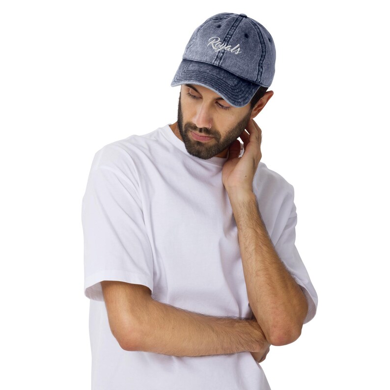 May include: A denim blue baseball cap with the word "Royals" embroidered in white script. The cap has a curved brim and a distressed, vintage look. The cap is worn by a person wearing a white t-shirt.