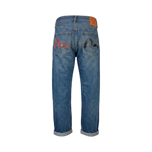 May include: Blue denim jeans with red and black graphic designs on the back pockets. The jeans have a classic five-pocket design, a leather patch, and a relaxed fit. The cuffs are rolled up.