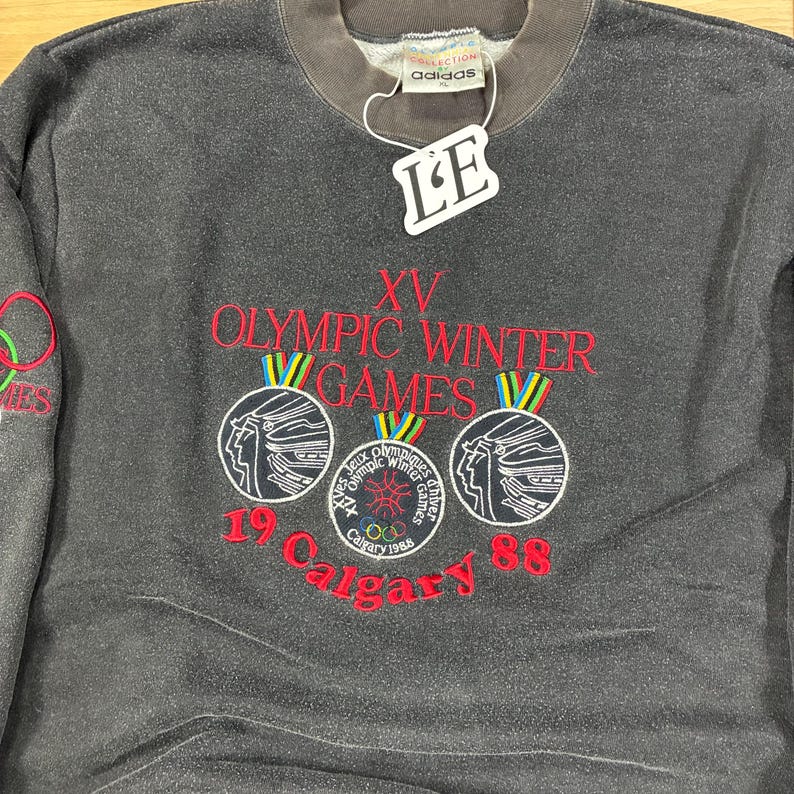 May include: Dark gray Adidas sweatshirt with red and white embroidered text and graphics. The text reads "XV Olympic Winter Games Calgary 88". Features Olympic rings and a tag that says "LE".