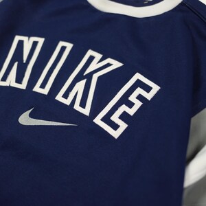 Op de afbeelding: Marineblauw sportshirt met het woord "NIKE" in witte blokletters en een grijs Nike swoosh-logo. Het shirt heeft een witte kraag en grijs-witte mouwen. Het shirt is gemaakt van ademend materiaal.