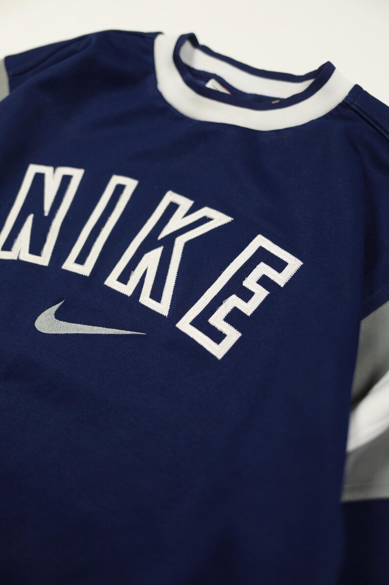 Op de afbeelding: Marineblauw sportshirt met het woord "NIKE" in witte blokletters en een grijs Nike swoosh-logo. Het shirt heeft een witte kraag en grijs-witte mouwen. Het shirt is gemaakt van ademend materiaal.