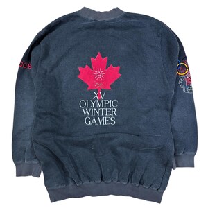 May include: A vintage dark gray sweatshirt with the text "XV Olympic Winter Games" and a red maple leaf emblem. The sweatshirt also features the Adidas logo in red and a colorful Olympic rings design.