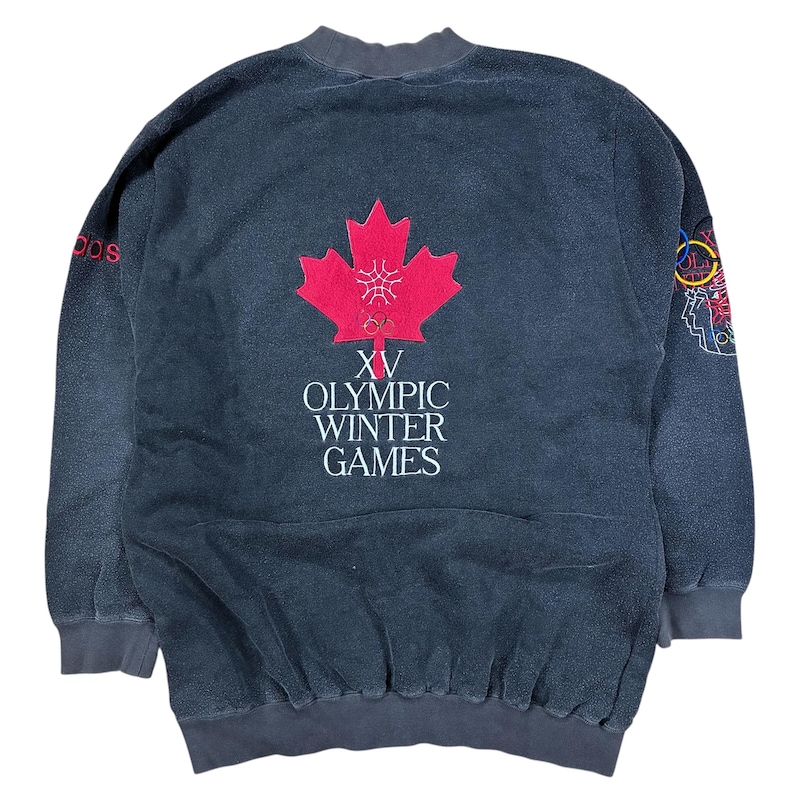 May include: A vintage dark gray sweatshirt with the text "XV Olympic Winter Games" and a red maple leaf emblem. The sweatshirt also features the Adidas logo in red and a colorful Olympic rings design.