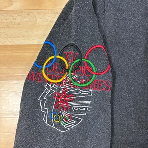 May include: Dark gray sweatshirt sleeve with embroidered Olympic rings in blue, yellow, black, green, and red. The text "XV Olympic Winter Games" is embroidered in red and white, along with a white outline of a person's face and other Olympic symbols.