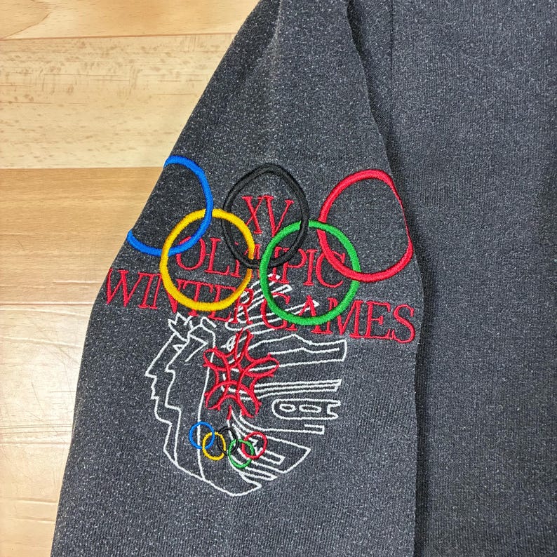 May include: Dark gray sweatshirt sleeve with embroidered Olympic rings in blue, yellow, black, green, and red. The text "XV Olympic Winter Games" is embroidered in red and white, along with a white outline of a person's face and other Olympic symbols.