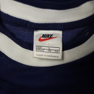 Op de afbeelding: Close-up van een marineblauw sportshirt met een wit en marineblauwe gestreepte kraag. Het Nike-logo is zichtbaar op een label, samen met informatie over maat en lengte. Het label geeft ook aan dat het shirt is gemaakt in Indonesi&euml;.