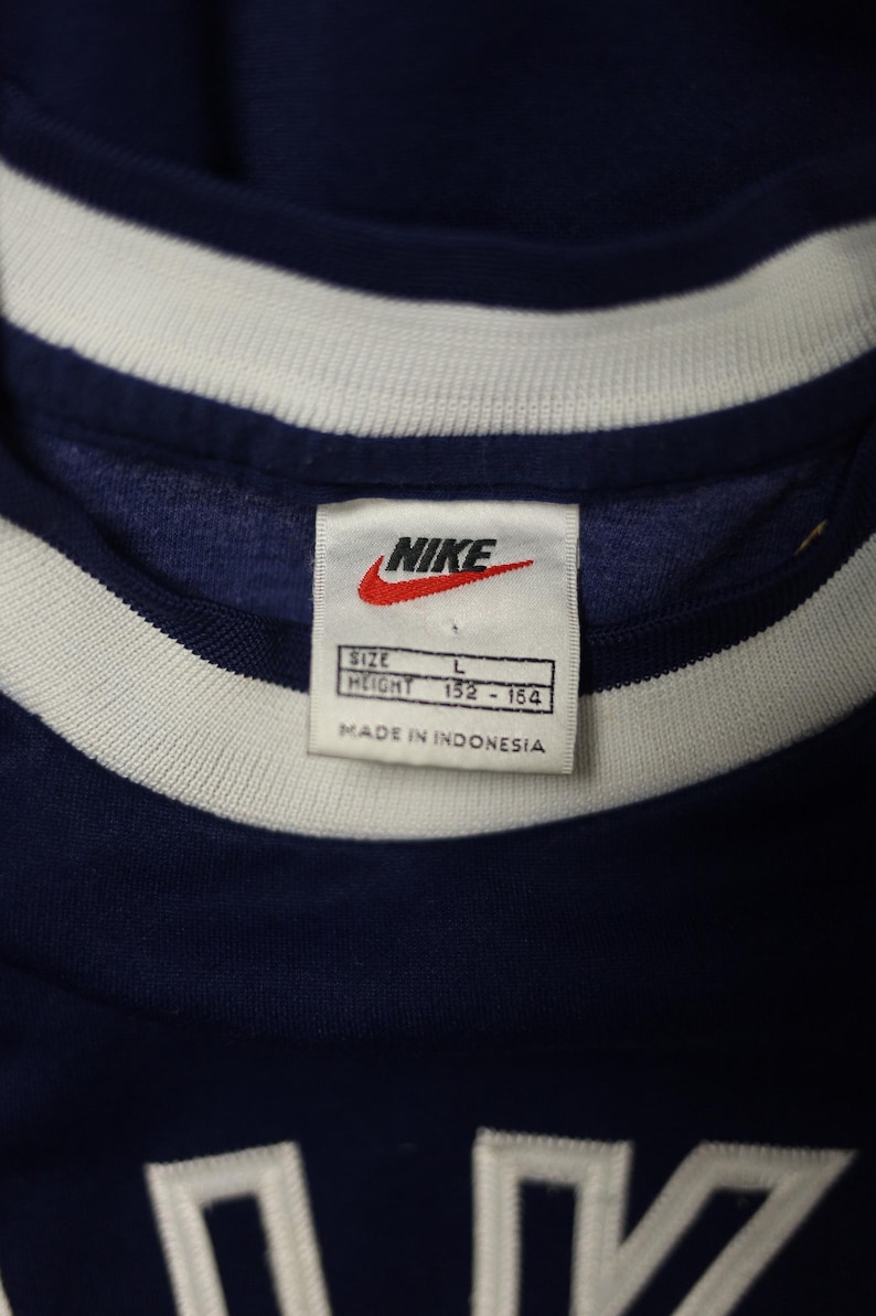 Op de afbeelding: Close-up van een marineblauw sportshirt met een wit en marineblauwe gestreepte kraag. Het Nike-logo is zichtbaar op een label, samen met informatie over maat en lengte. Het label geeft ook aan dat het shirt is gemaakt in Indonesi&euml;.
