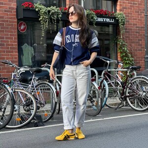 Op de afbeelding: Een persoon draagt een marineblauw sweatshirt met "NIK" op de voorkant, gecombineerd met een lichte jeans en gele sneakers. De persoon staat op een straat met fietsen op de achtergrond, voor een bakstenen gebouw.