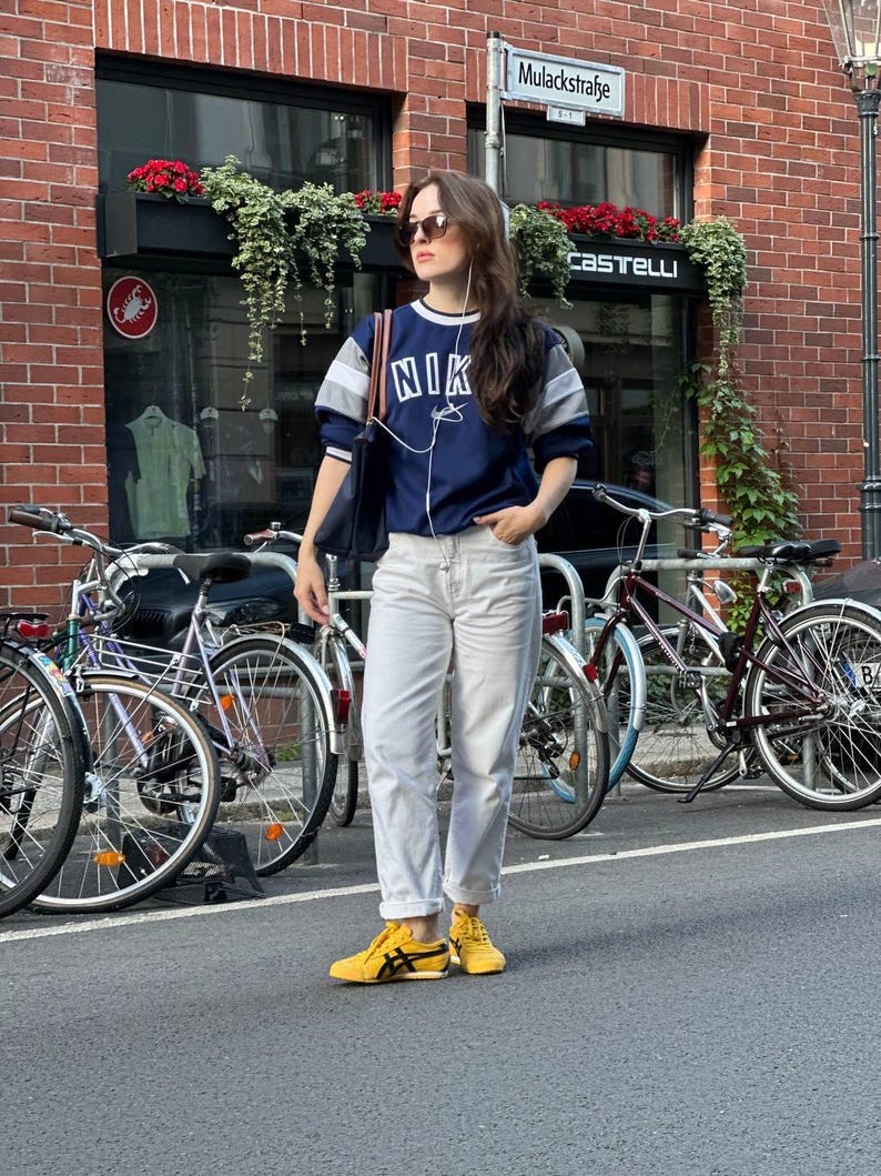 Op de afbeelding: Een persoon draagt een marineblauw sweatshirt met "NIK" op de voorkant, gecombineerd met een lichte jeans en gele sneakers. De persoon staat op een straat met fietsen op de achtergrond, voor een bakstenen gebouw.